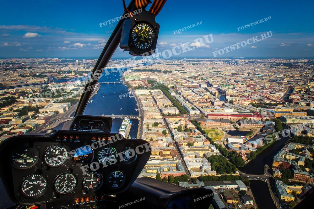 Фотография демонстрирует вид на Санкт-Петербург из кабины вертолёта, создавая эффект присутствия и впечатление личного полёта над городом. Под крылом вертолёта простирается величественная река Нева, омывающая исторические здания в центре города. Прямоугольные улицы и кварталы гармонично сочетаются с извилистым течением реки, а множество деталей позволяет рассматривать городскую структуру. Эта фотография может быть использована для тематических публикаций о полётах, городском планировании и туристических презентаций Санкт-Петербурга.